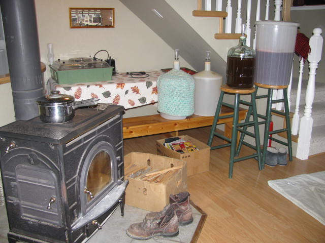A woodstove burning with a pot cooking on top. Work boots in front drying, and carboys of wine being made.