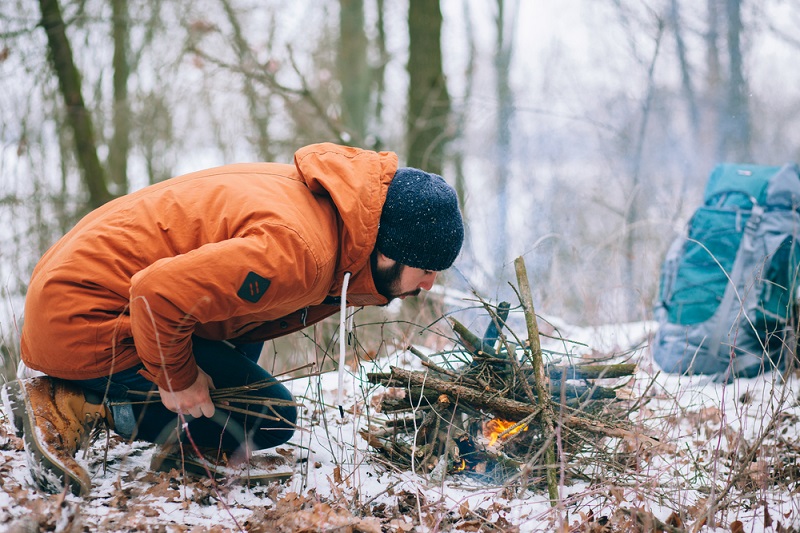 a bundled up man lights a campfire in winter