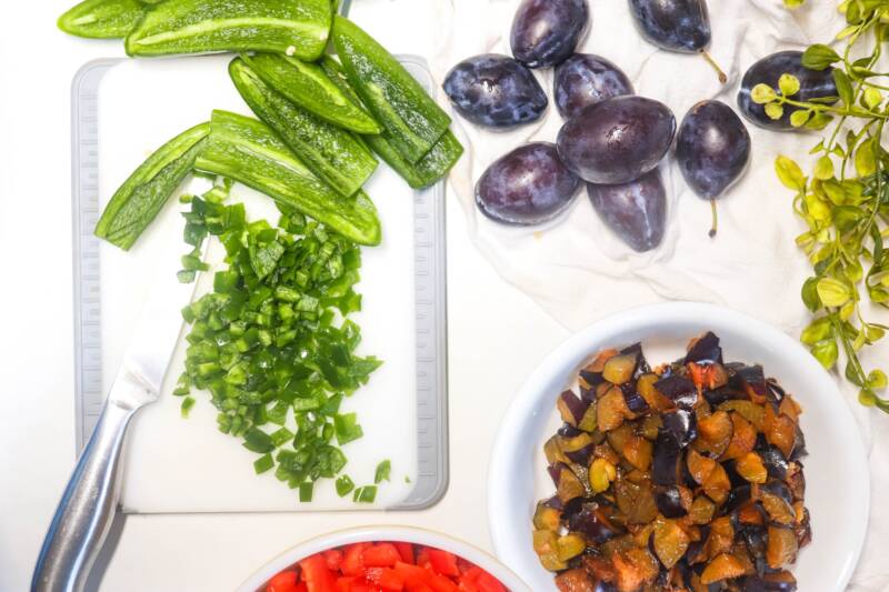 Diced Jalapeno and red and green peppers on a cutting board along with plums.
