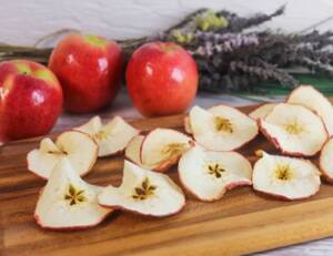 Dehydrated apples sit on a wood tray with whole apples and lavender behind.